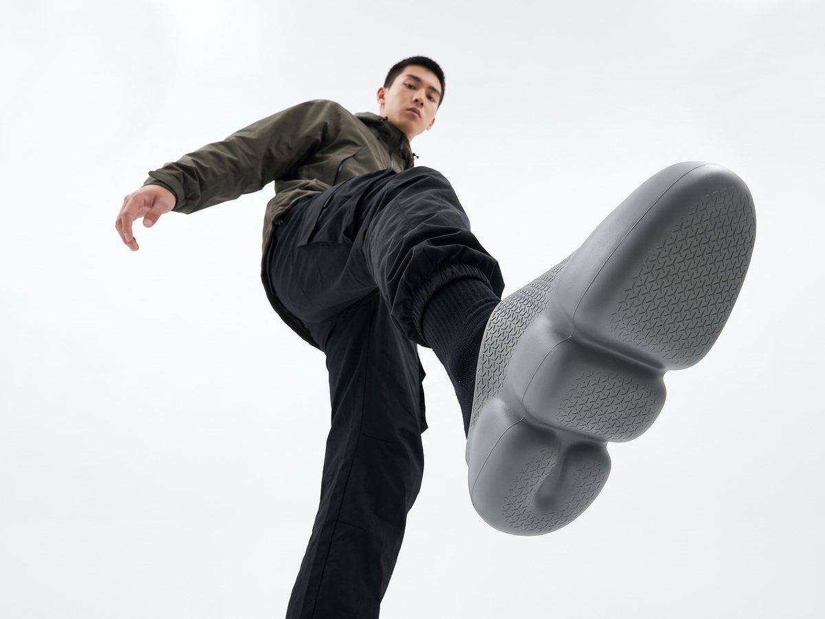 Seen from below, a model in black kicks toward the camera, the sole of the grey Kimi mule filling the frame
