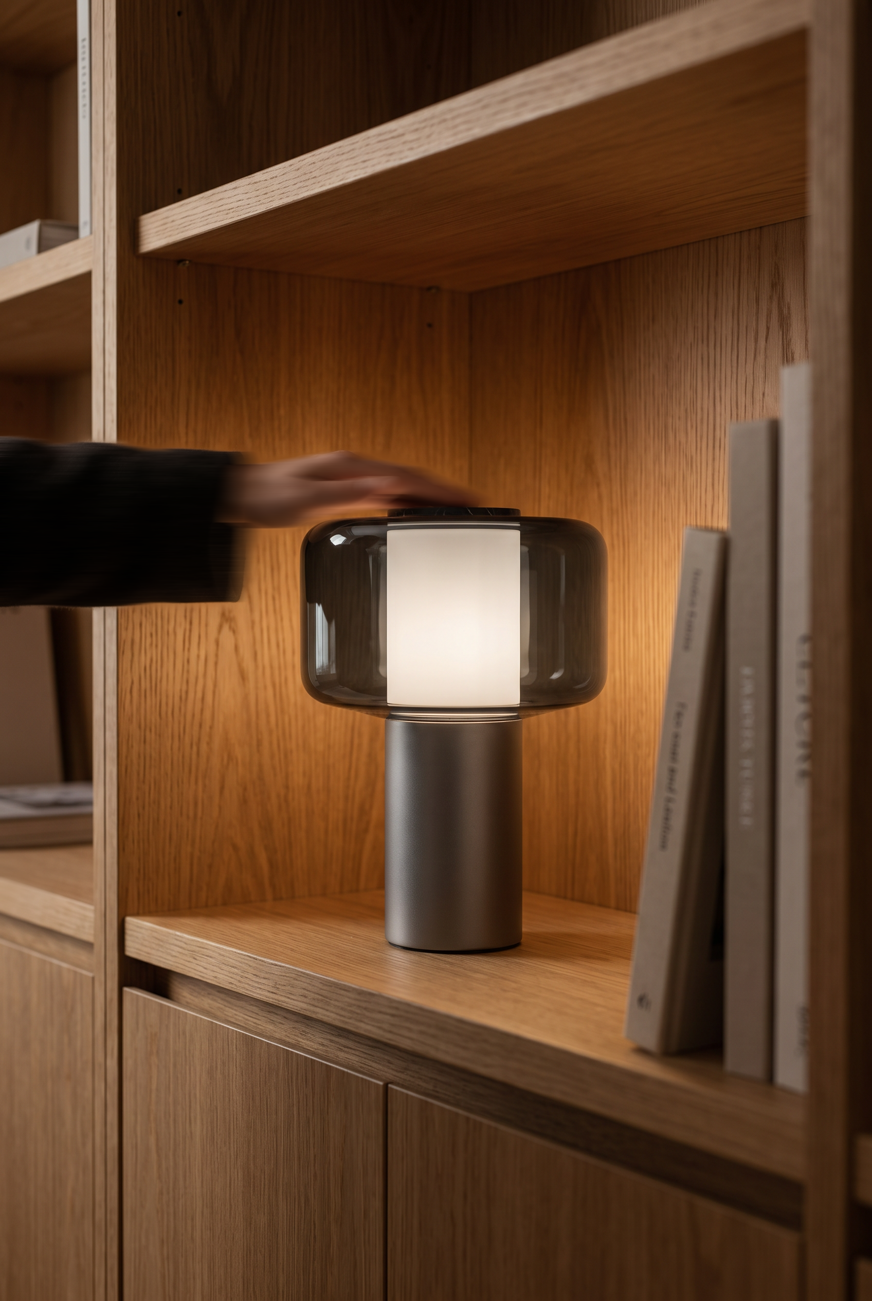 A hand reaches to the marble cap of Lumen, lit on an oak bookshelf, the arm blurred in motion